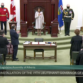 A woman dressed in white stands in front of a wooden throne flanked by three military members. She is facing a room full of applauding audience members who are facing her.