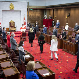 A procession of people are walking along a red carpet towards the front of the room. Three of the individuals are wearing black. One woman is wearing white. There is a throne chair flanked by two Canada flags. 