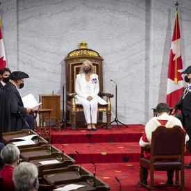 A woman dressed in a white suit is sitting on a throne chair. There are Canada flags on either side of the platform. There is a man sitting in a chair facing her. He is wearing a red and white cape.