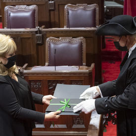 A man and a woman are both dressed in black, wearing masks. They are both holding a binder that has a green bow on it. The man is wearing white gloves and a black hat.