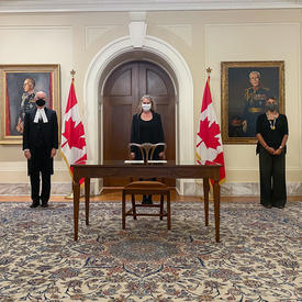 Quatre personnes portant des masques faciaux se tiennent au fond de la salle. Il y a une table et une chaise en bois devant la femme au centre de la photo. Il y a deux drapeaux canadiens de chaque côté d'elle.