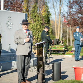 Mr. Whit Grant Fraser is standing at a podium. The podium features the badge of the Canadian Forces. He is wearing a poppy. Five people wearing a military uniform are standing behind him. 