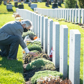 Mr. Whit Grant Fraser is kneeling by a headstone at the Beechwood Cemetery. He is placing a poppy at the foot of the headstone. He is wearing a mask. It is sunny.