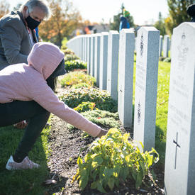 A student is kneeling in front of a headstone and is placing a poppy at its base. Mr. Whit Grant Fraser is crouching next to her, looking at the poppy. He is wearing a mask. There are many rows of headstones in the background.