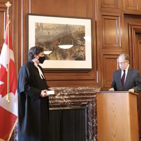 Chief Justice Richard Wagner holding a book. Ian Shugart - clerk of the Privy Council of Canada - stands a podium. 