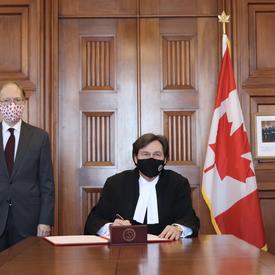 Chief Justice Richard Wagner sitting at a table. Ian Shugart - clerk of the Privy Council of Canada - standing to his right.