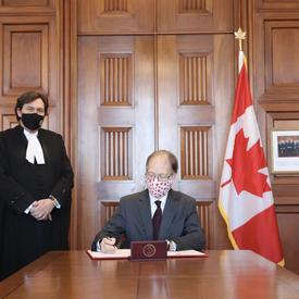 Ian Shugart - clerk of the Privy Council of Canada - sitting at a table. Chief Justice Richard Wagner standing to his right.