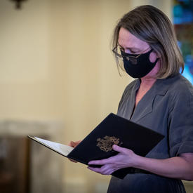 A woman reading from a document. She is wearing a black mask.