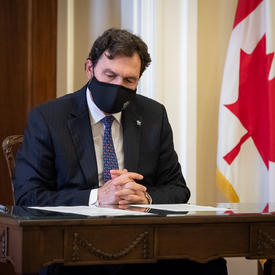 The Administrator reading a document while seated at a table. There is a Canadian flag in the background.