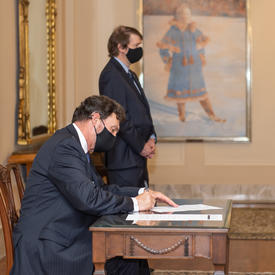 L’administrateur, assis à une table, signe un document. On aperçoit le secrétaire à l’arrière-plan.