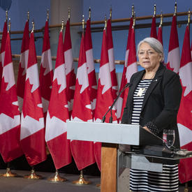 Prime Minister Justin Trudeau and Governor General Designate Mary May Simon each stand at a podium with several Canadian flags behind them. Mary Simon is speaking. Justin Trudeau's head is turned towards her.