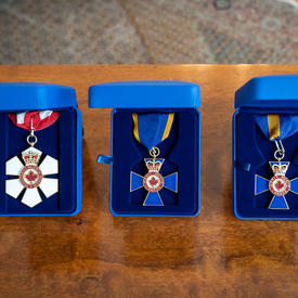 Three Canadian order decorations are positioned on a wood table in individual blue suede boxes. The first decoration is red and white with a red and white ribbon. The second and third decorations are blue with blue ribbon with a yellow border. 