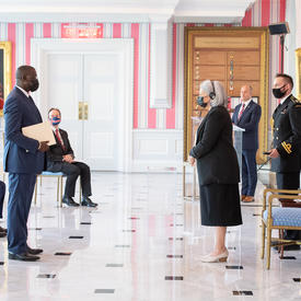 A man in a suit and a face mask hands a document to the Governor General.