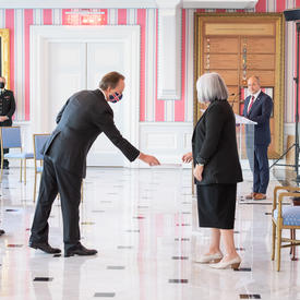 A man in a suit and a face mask hands a document to the Governor General.