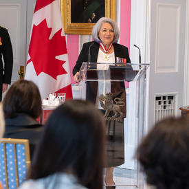 La gouverneure générale Mary May Simon, debout derrière un lutrin en verre, s’adresse au public rassemblé dans la salle de la tente de RideauLa maîtresse de cérémonie annonce le nom des récipiendaires d’une distinction honorifique debout devant le lutrin.