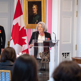 Governor General Mary May Simon stands at a glass podium addressing the audience in the Tent Room at Rideau Hall. There is a large Canadian flag behind her.