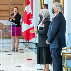 La gouverneure générale Mary May Simon applaudit. Son Excellence Whit Fraser est debout, les mains croisées devant lui. Un drapeau canadien et la maîtresse de cérémonie sont en arrière-plan.