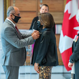 A man pins an award to a woman’s jacket, with Governor General Mary May Simon looking on. A Canadian flag is in the background.