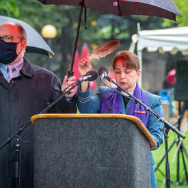 A woman stands at a podium. A man holds an umbrella over her to protect her against the rain.
