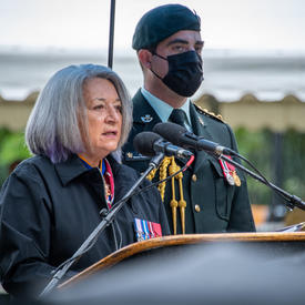 The Governor General gives her speech. She is outdoors. A man in military uniform stands behind her, wearing a mask. It is raining.
