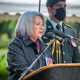 La gouverneure générale prononce son allocution. Un homme en uniforme militaire portant un masque se tient derrière elle. Il pleut. 