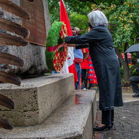 The Governor General lays a wreath at the base of the National Aboriginal Veterans Monument. Feathers are visible on the left side of the photo. In the background are trees and a small crowd of people wearing masks.