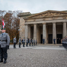 Large building in the background. Members of the German military are standing in uniform. They are outdoors.