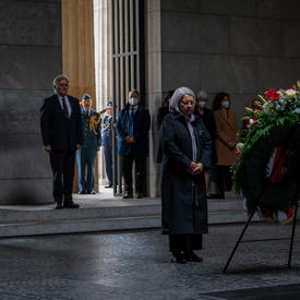 Governor General Mary May Simon is standing in front of a large wreath. Her hands are joined in front of her. There are people standing along the wall behind her.