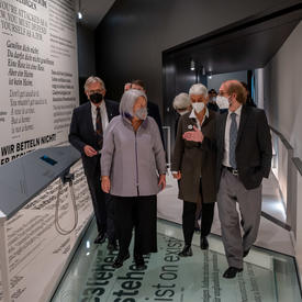  Their Excellencies are walking down the hallway in a museum. A small group of people are following them. 