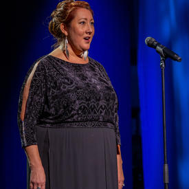 A lady is singing. She is standing at a microphone. She is dressed in formal evening wear. 