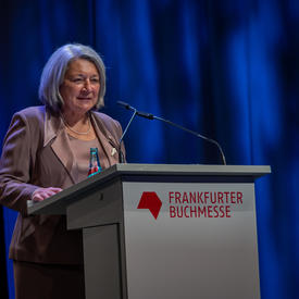 Governor General Mary May Simon is standing behind a grey podium. She is wearing a rose pink suit. Behind her is a dark blue curtain. On the podium, there is red text that reads, “Frankfurter Buchmesse.” 
