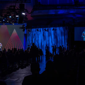 Her Excellency is delivery remarks at a podium. The room is dark and there is a light show which surrounds her. 