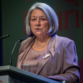 Governor General Mary May Simon is standing behind a grey podium. She is wearing a rose pink suit. Behind her is a dark blue curtain.