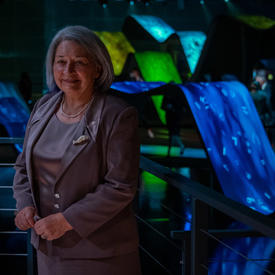Governor General Mary May Simon is standing in front of a large illuminated art display made of blue, green, and yellow ramps with projections on them. She is smiling.