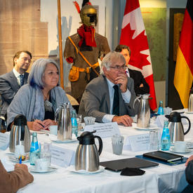 The Governor General and Mr. Fraser are sitting at a table with six other people. Ms. Simon is looking down at the table and Mr. Fraser is speaking while gesturing with his hands. 