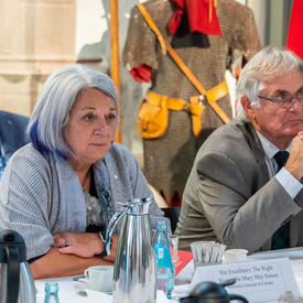 The Governor General and Mr. Fraser are sitting at a table with six other people. Ms. Simon is looking down at the table and Mr. Fraser is speaking while gesturing with his hands. 