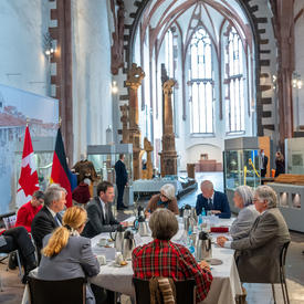 The Governor General and Mr. Fraser are sitting at a table with six other people. In the background there is a Canadian flag and to its left a German flag. There is also a suit of armour on a display rack.