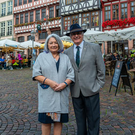 Governor General Mary May Simon and Mr. Fraser pose on a street in Old Town Frankfurt. Mr. Fraser has his arm around Ms. May Simon and they are smiling. There are colourful buildings in the background and several vendor tents.