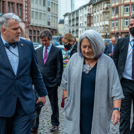 Governor General Mary May Simon on a street in Old Town Frankfurt. She is beside a man who is accompanying her. There are colourful buildings in the background and several vendor tents.uildings in the background and several vendor tents.