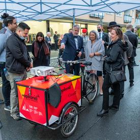 Their Excellencies are outdoors. They are standing with a group of people admiring a bicycle cart from the Frankfurt Book Fair. 