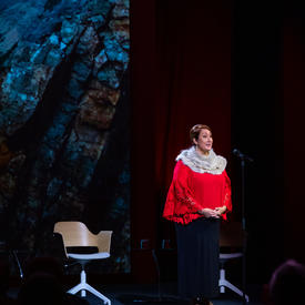 Deantha Edmunds is standing on a stage with her hands clasped in front of her. She is wearing a red shirt with white fur around the collar and a navy skirt. She is speaking into a microphone. 