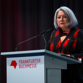 Governor General Mary May Simon is standing behind a grey podium. She is speaking into a microphone. On the podium, there is red text that reads, “Frankfurter Buchmesse.” She is wearing a black and red shirt.