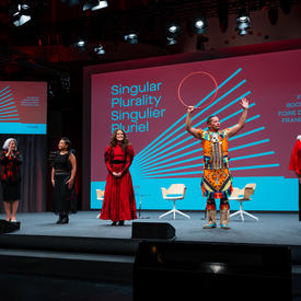 Governor General Mary May Simon is standing on a stage. To her right is a grey podium, to her left are four performers, in order an accompanying musician, Iskwē, Dallas Arcand, and Deantha Edmunds. She is looking at them and clapping. 