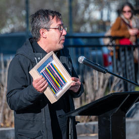 A man speaks at a podium. He is holding a photo box that has a row of crayons in it.