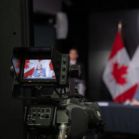 A camera is filming Governor General Mary Simon. She can be seen on the LCD screen of the camera. She is also in the background of the photo, out of focus. Behind her are Canadian flags.