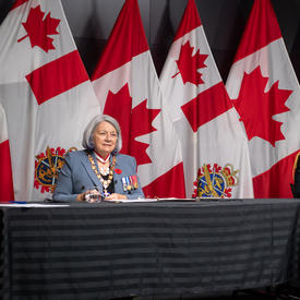 La gouverneure générale Mary Simon est assise à une longue table avec le général Wayne Eyre. Ils font tous deux face à la caméra. Derrière eux il y a des drapeaux canadiens et des Forces armées canadiennes.