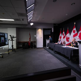 Governor General Mary Simon is sitting at a long table with General Wayne Eyre. There are people in military uniform standing at podiums on either side of the table. 