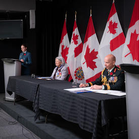 Governor General Mary Simon is sitting at a long table with General Wayne Eyre. There are people in military uniform standing at podiums on either side of the table. 