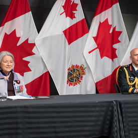La gouverneure générale Mary Simon est assise à une longue table avec le général Wayne Eyre. Ils font tous deux face à la caméra. Derrière eux il y a des drapeaux canadiens et des Forces armées canadiennes.