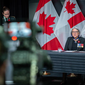 Une caméra enregistre la gouverneure générale Mary Simon. On la voit sur l'écran LCD de la caméra. Elle est également à l'arrière-plan de la photo. Derrière elle, on aperçoit des drapeaux canadiens.